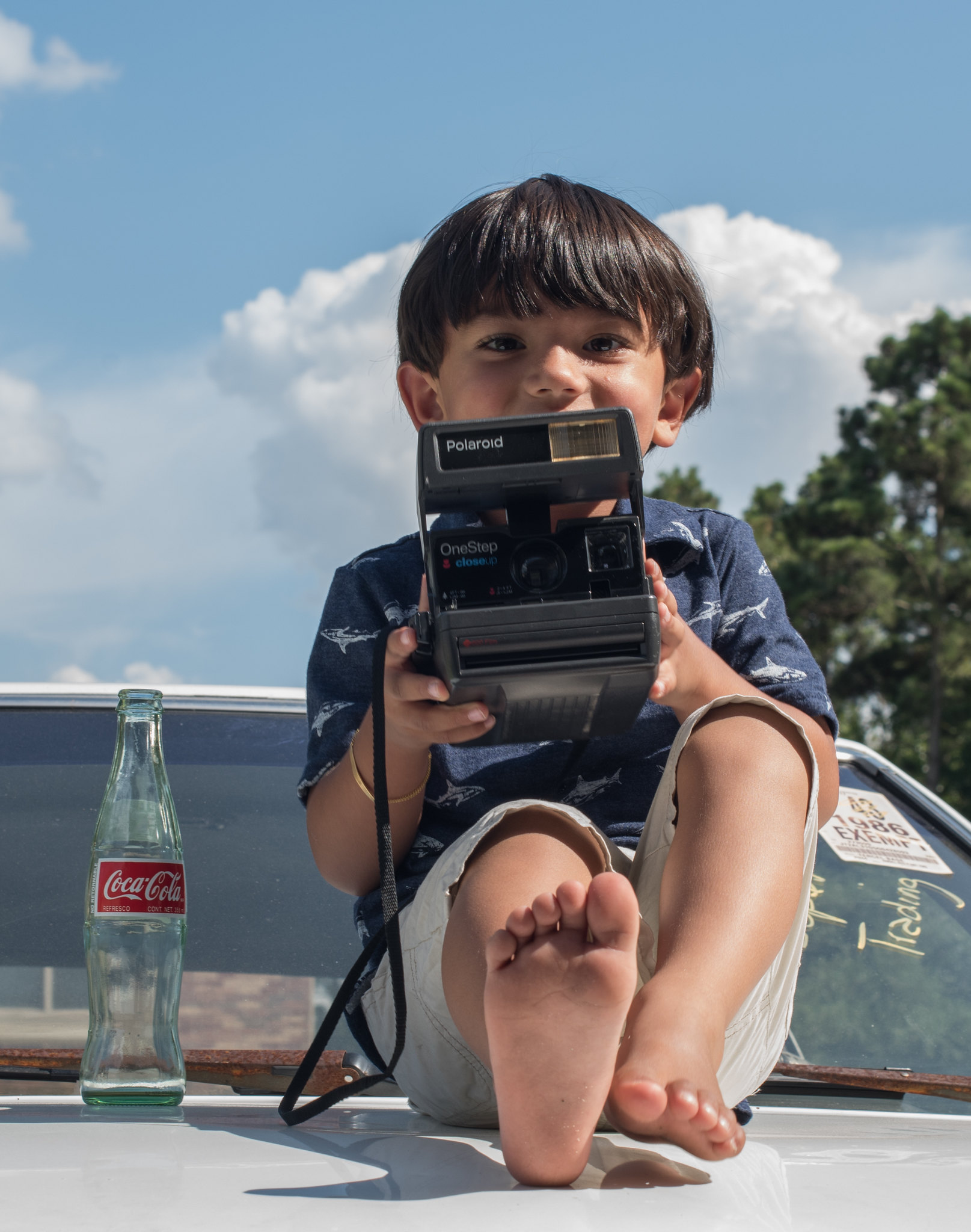 A boy with a Polaroid camera and a glass Coca-Cola bottle, sitting on a car hood.